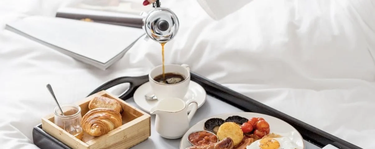 Breakfast tray on a hotel bed with coffee being poured, croissants, fruit and preserves