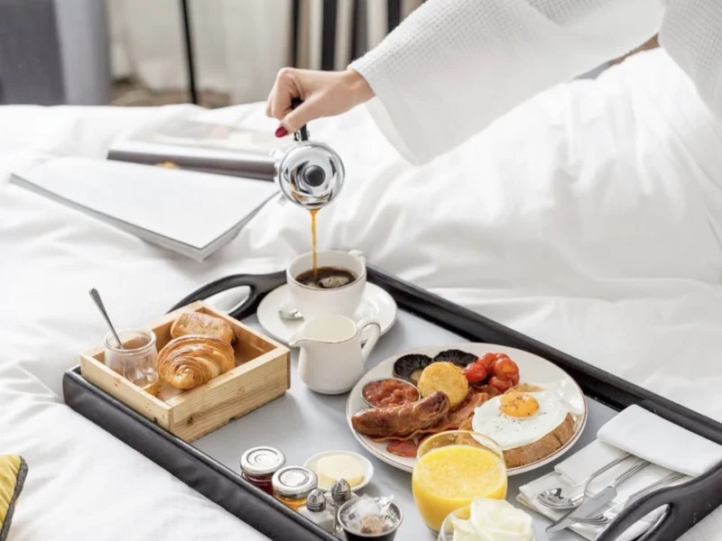 Breakfast tray on a hotel bed with coffee being poured, croissants, fruit and preserves