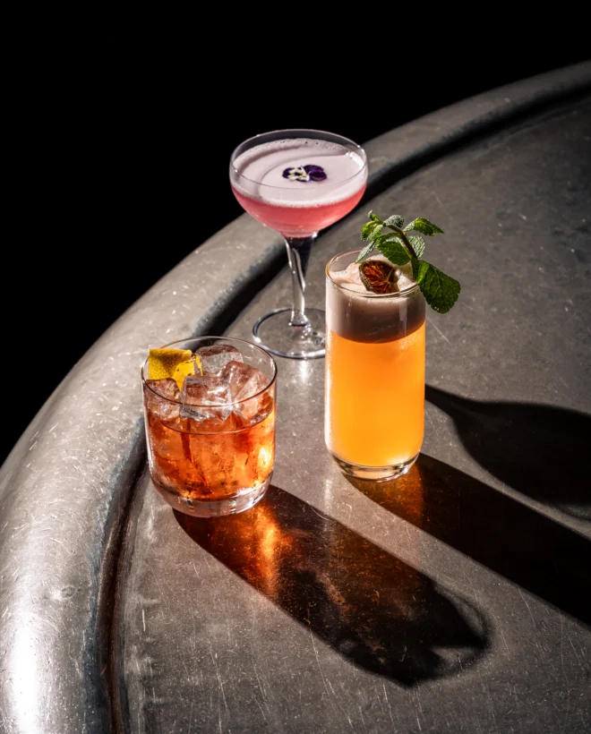 Two cocktails on a bar counter, a pink coupe and an amber highball, in low light