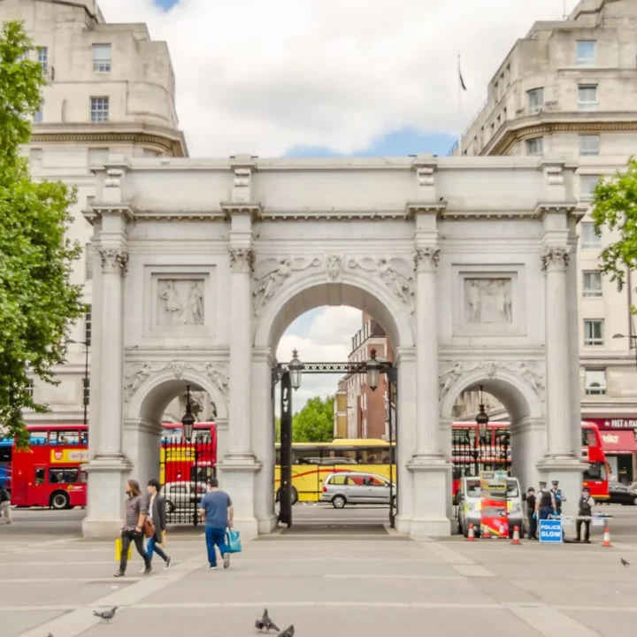Marble Arch landmark in London with red double-decker buses, international flags, pedestrians, and green trees.