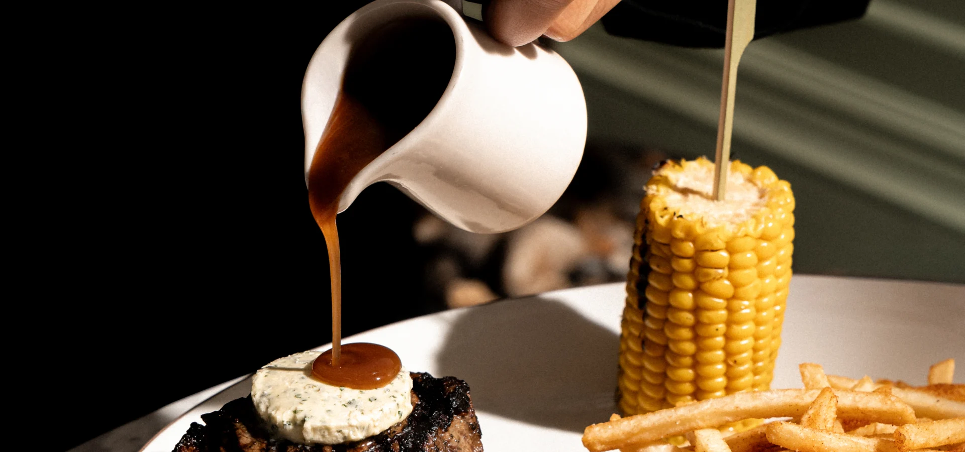 Peppercorn sauce being poured over steak with fries, onion rings and corn
