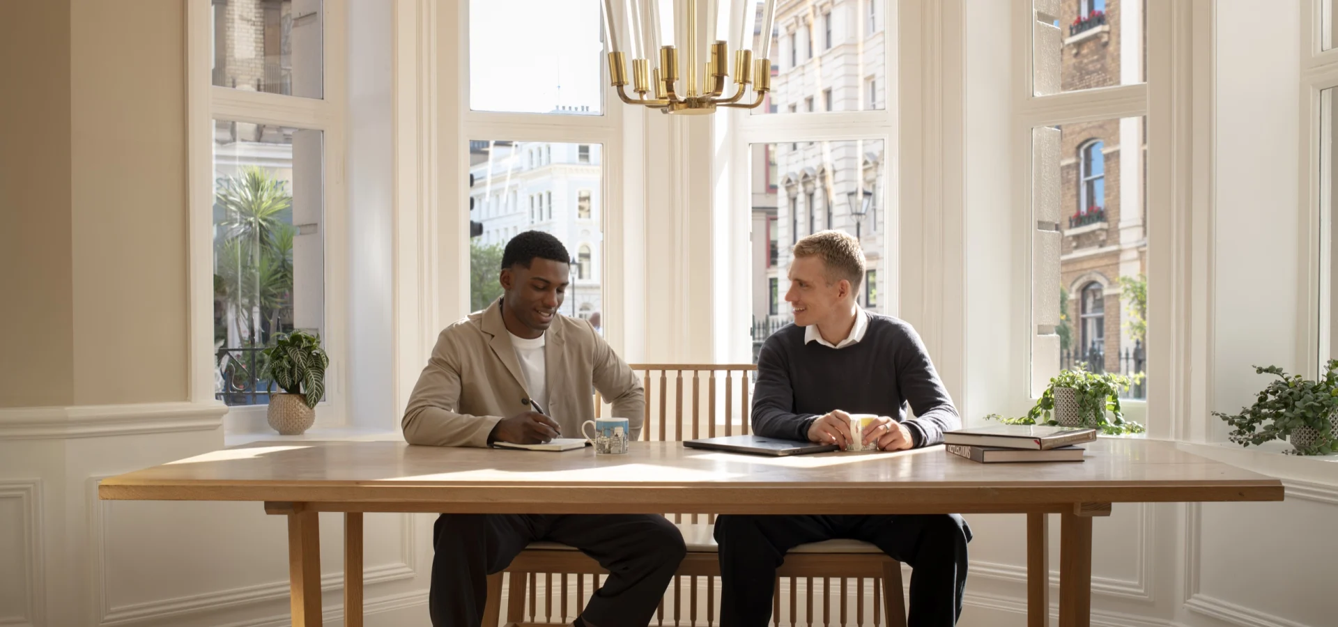 Two men meeting at a wooden table beside a bay window at Guild Hotel Bloomsbury