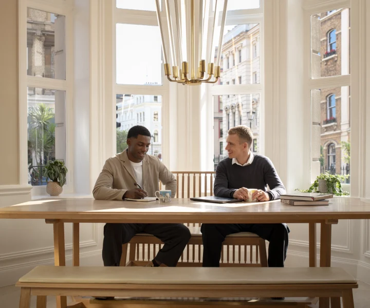 Two men meeting at a wooden table beside a bay window at Guild Hotel Bloomsbury