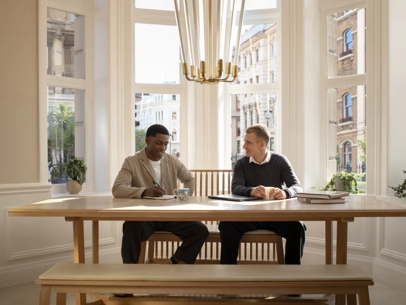 Two men meeting at a wooden table beside a bay window at Guild Hotel Bloomsbury