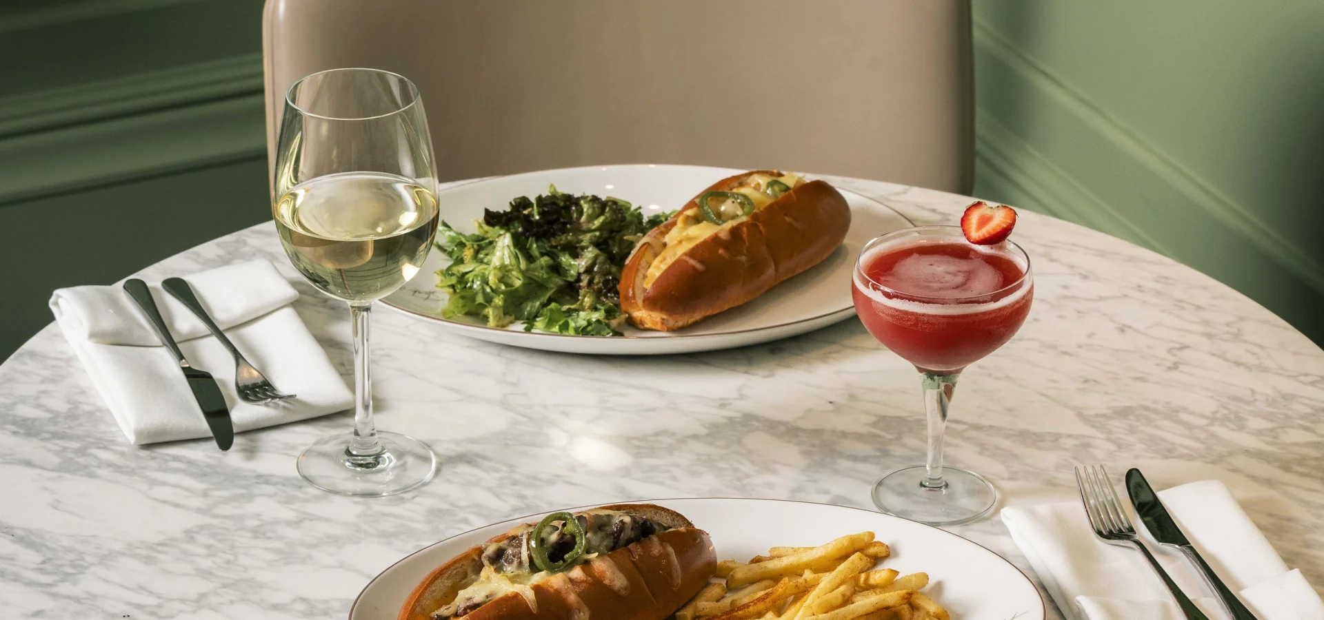 Sandwiches, fries, salad, wine and a cocktail on a marble table for two