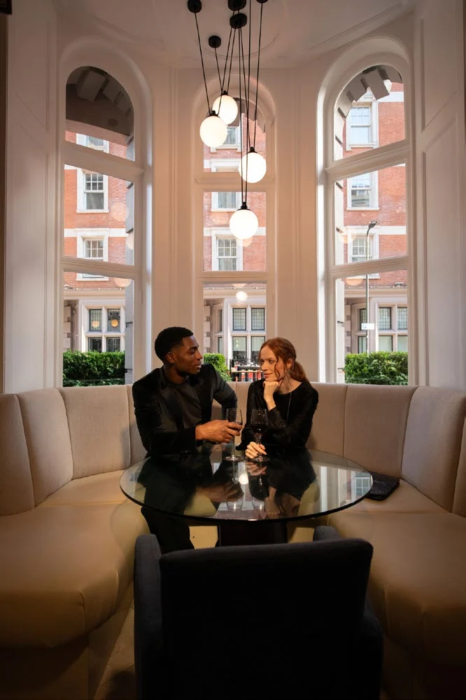 Couple with drinks on a curved banquette beneath globe pendant lights in the bay window of the Bloomsbury Street Lounge at dusk