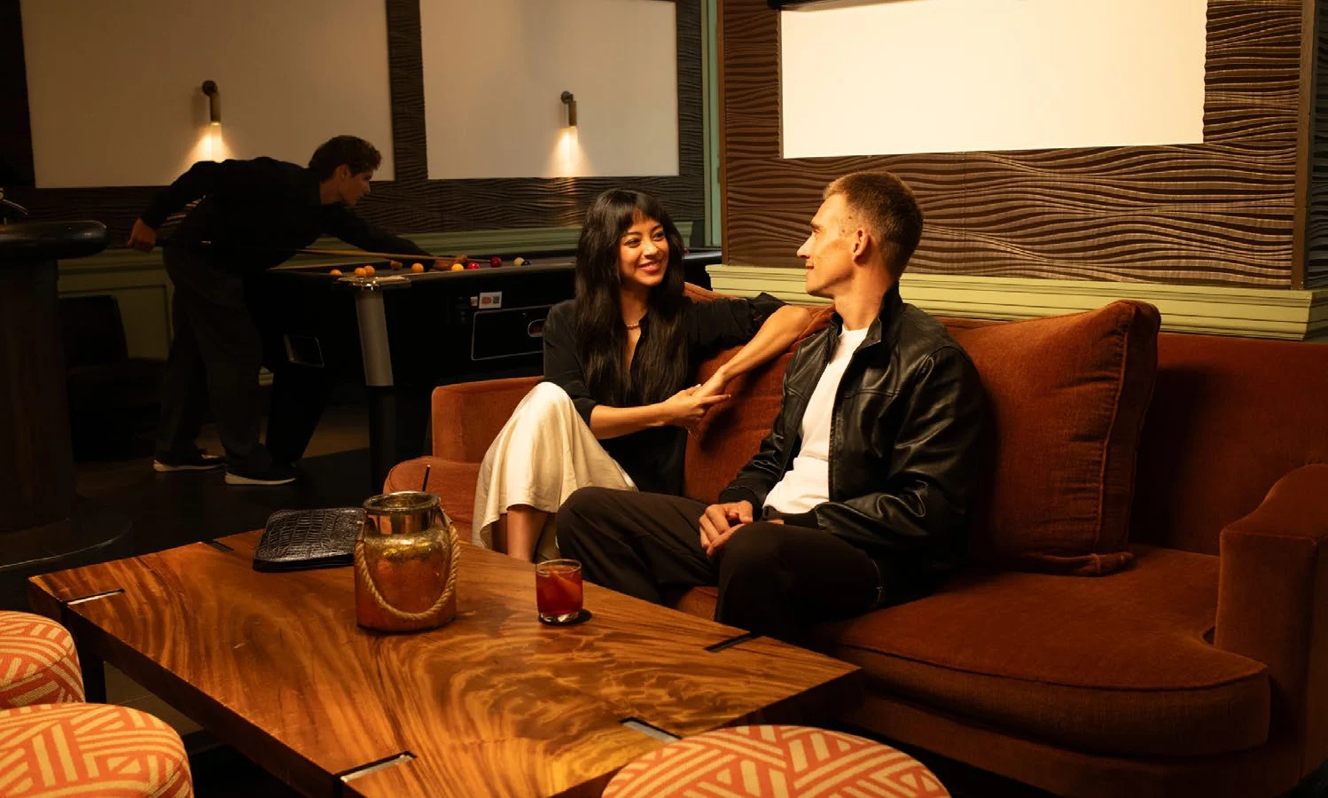 Couple on a rust velvet sofa in The Warren, pool table and sports screens behind